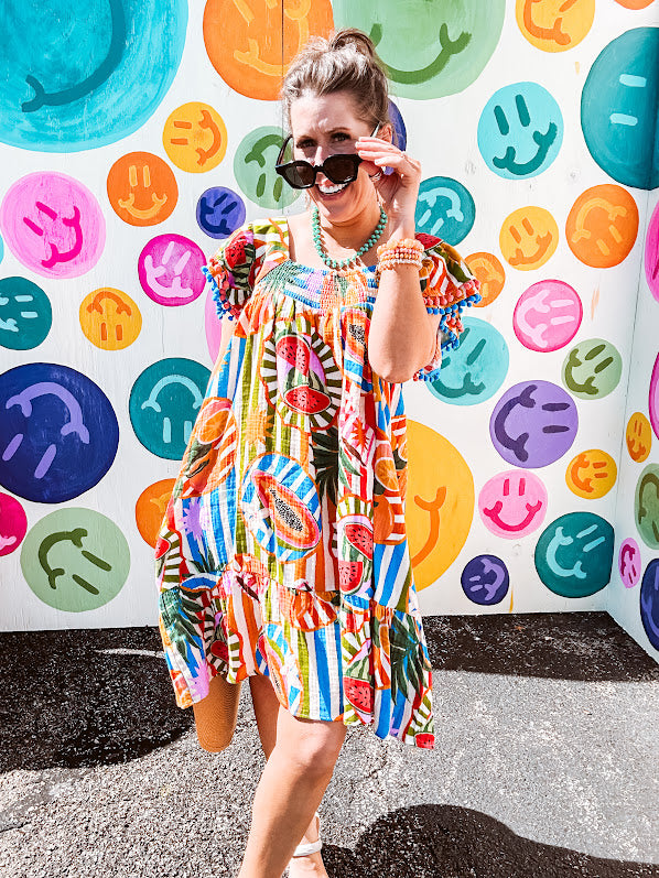 This tropical print short dress features vibrant summer hues with playful striping and fruit motifs throughout, square smocked neckline and flutter sleeves and pockets—complete with pink and blue ball tassels. Crafted from soft, lightweight fabric, it's designed to move with you all day long.