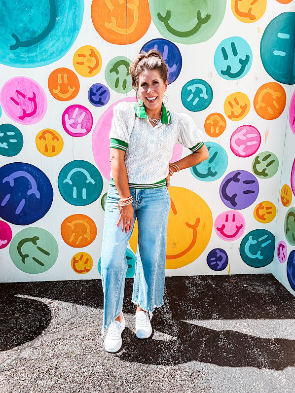 Nearly sheer ivory eyelet patterned blouse with a collar, puff sleeves and three large rhinestone floral buttons at the neckline. The collar, cuffs and hem are a contrasting bright green with pink athletic striping.