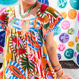 This tropical print short dress features vibrant summer hues with playful striping and fruit motifs throughout, square smocked neckline and flutter sleeves and pockets—complete with pink and blue ball tassels. Crafted from soft, lightweight fabric, it's designed to move with you all day long.