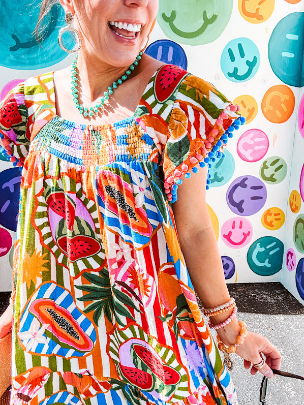 This tropical print short dress features vibrant summer hues with playful striping and fruit motifs throughout, square smocked neckline and flutter sleeves and pockets—complete with pink and blue ball tassels. Crafted from soft, lightweight fabric, it's designed to move with you all day long.