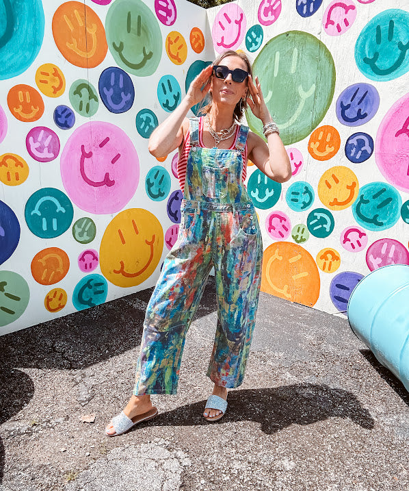 Cotton denim overalls with adjustable straps and sides with metal buttons in a paint splatter detail in cobalt blue, orange, green and yellow washed color with knee darts.