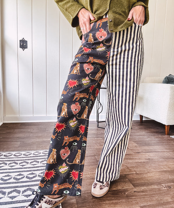 Lightweight denim-like non-stretch wide leg pants with elastic back waist, side and back pockets, button and zipper closure and black and cream stripes on one leg and funky leopard, heart, eye graphics on a black background on the other.