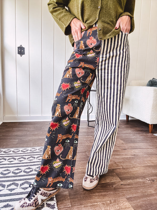 Lightweight denim-like non-stretch wide leg pants with elastic back waist, side and back pockets, button and zipper closure and black and cream stripes on one leg and funky leopard, heart, eye graphics on a black background on the other.
