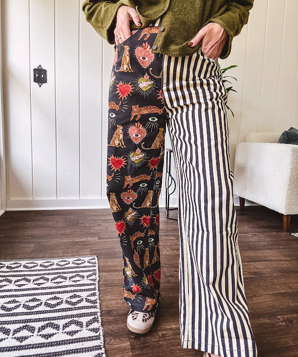 Lightweight denim-like non-stretch wide leg pants with elastic back waist, side and back pockets, button and zipper closure and black and cream stripes on one leg and funky leopard, heart, eye graphics on a black background on the other.