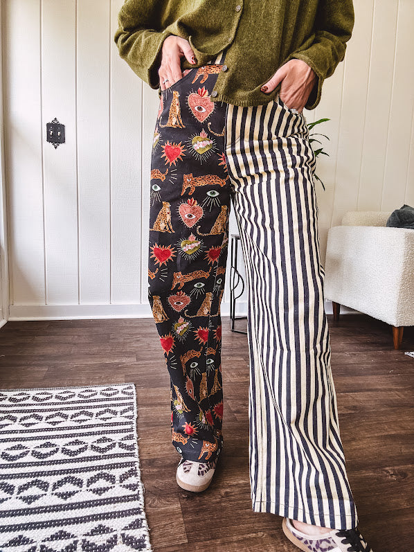 Lightweight denim-like non-stretch wide leg pants with elastic back waist, side and back pockets, button and zipper closure and black and cream stripes on one leg and funky leopard, heart, eye graphics on a black background on the other.