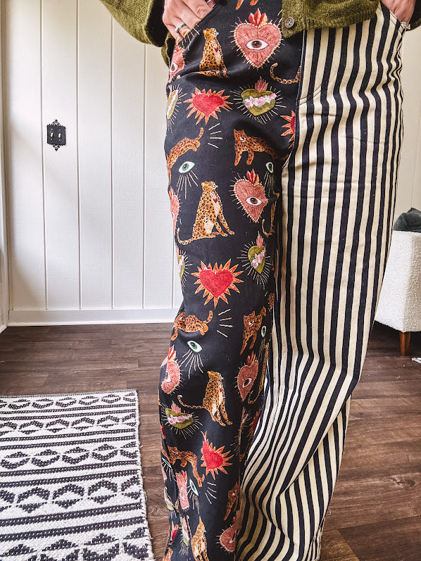 Lightweight denim-like non-stretch wide leg pants with elastic back waist, side and back pockets, button and zipper closure and black and cream stripes on one leg and funky leopard, heart, eye graphics on a black background on the other.