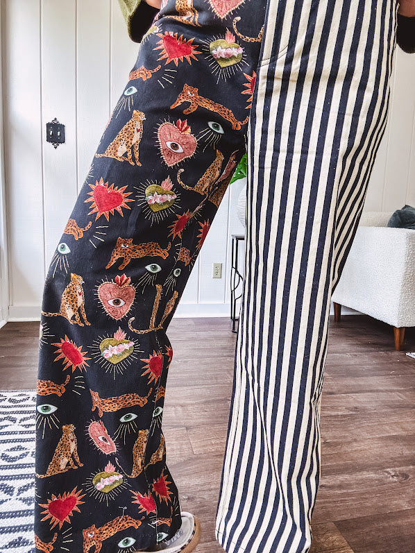 Lightweight denim-like non-stretch wide leg pants with elastic back waist, side and back pockets, button and zipper closure and black and cream stripes on one leg and funky leopard, heart, eye graphics on a black background on the other.