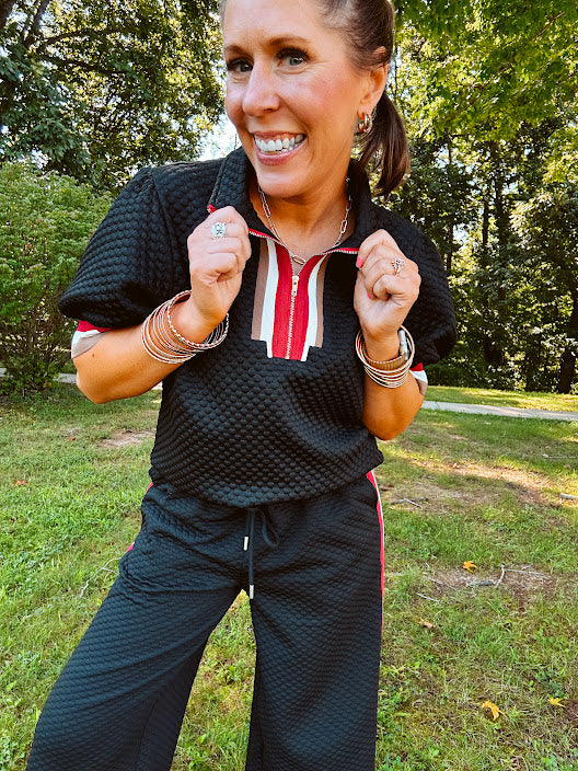 Black quilted short sleeved top with puff sleeves, 1/4 zip and taupe, red and cream striping detail at the neck and cuffs.