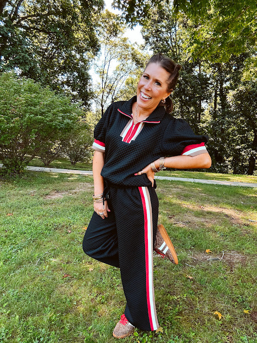 Black quilted short sleeved top with puff sleeves, 1/4 zip and taupe, red and cream striping detail at the neck and cuffs.