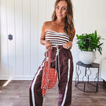 Dark brown slick track pants with white stripe detail down the sides and on the cutout hem with drawstring elastic waist and side pockets.