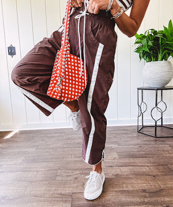Dark brown slick track pants with white stripe detail down the sides and on the cutout hem with drawstring elastic waist and side pockets.