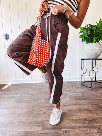 Dark brown slick track pants with white stripe detail down the sides and on the cutout hem with drawstring elastic waist and side pockets.