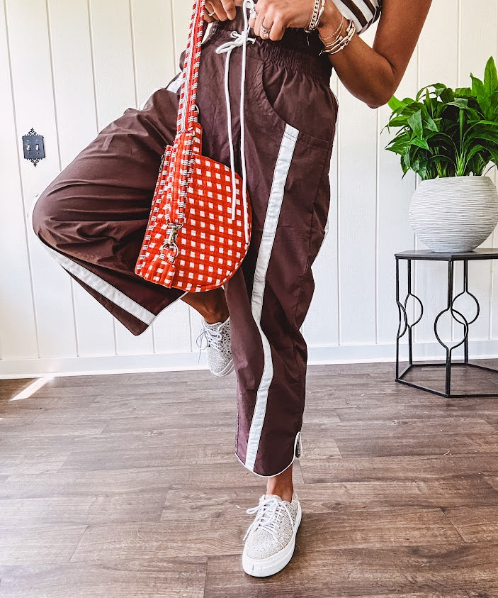 Dark brown slick track pants with white stripe detail down the sides and on the cutout hem with drawstring elastic waist and side pockets.
