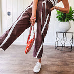 Dark brown slick track pants with white stripe detail down the sides and on the cutout hem with drawstring elastic waist and side pockets.