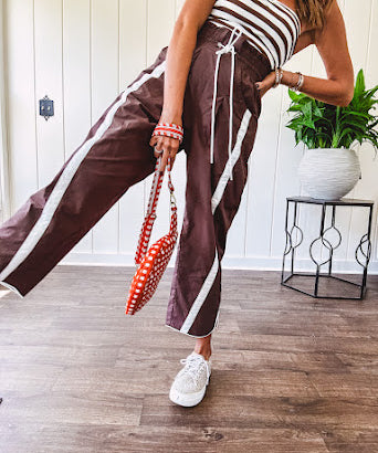 Dark brown slick track pants with white stripe detail down the sides and on the cutout hem with drawstring elastic waist and side pockets.