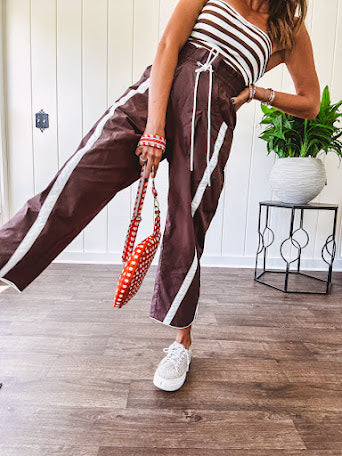 Dark brown slick track pants with white stripe detail down the sides and on the cutout hem with drawstring elastic waist and side pockets.
