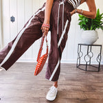 Dark brown slick track pants with white stripe detail down the sides and on the cutout hem with drawstring elastic waist and side pockets.