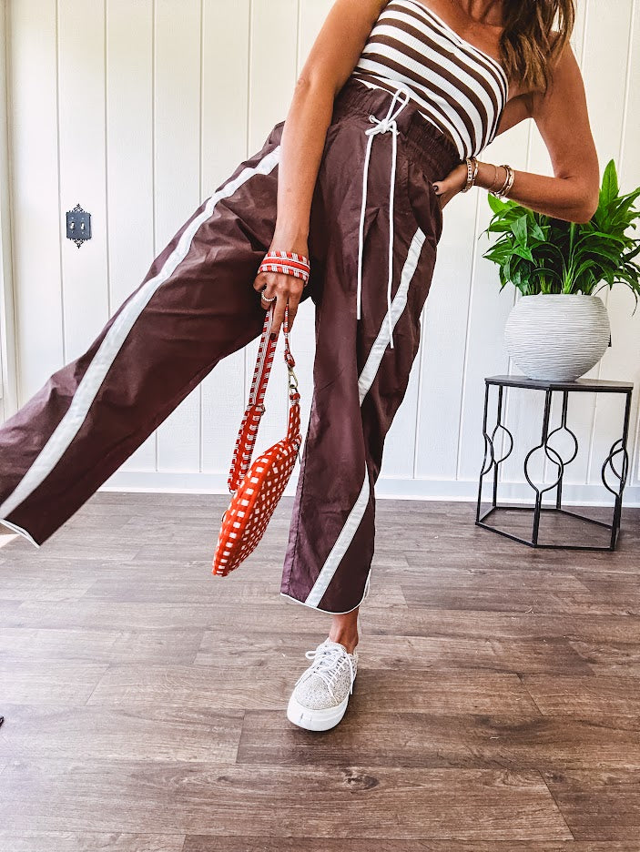 Dark brown slick track pants with white stripe detail down the sides and on the cutout hem with drawstring elastic waist and side pockets.
