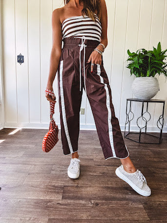 Dark brown slick track pants with white stripe detail down the sides and on the cutout hem with drawstring elastic waist and side pockets.