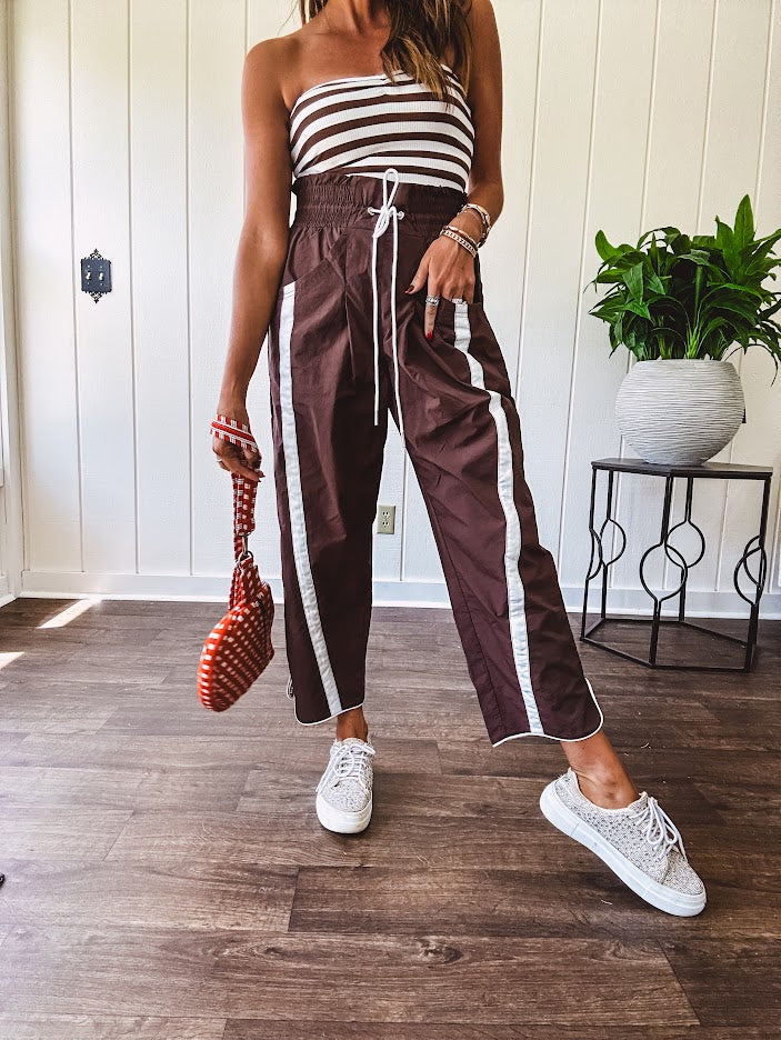 Dark brown slick track pants with white stripe detail down the sides and on the cutout hem with drawstring elastic waist and side pockets.