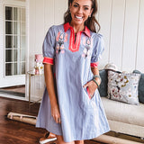 Slate blue dress with puffed, cuffed short sleeves, pockets, a contrasting coral collar and V neck, cuffs and trim and coral and pink embroidery at the yoke.