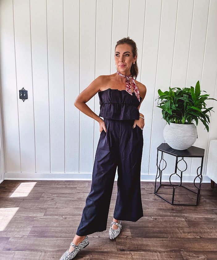 Fully lined black jumpsuit with sleeveless, ruffled bodice, smocked back, wide leg design and pockets at the pleated front.