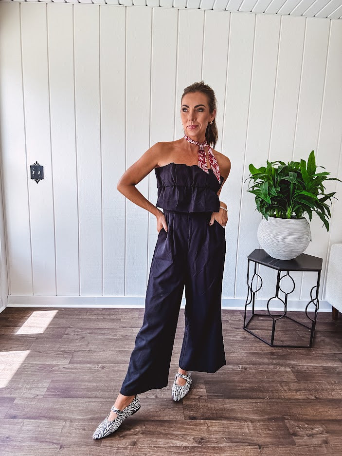 Fully lined black jumpsuit with sleeveless, ruffled bodice, smocked back, wide leg design and pockets at the pleated front.