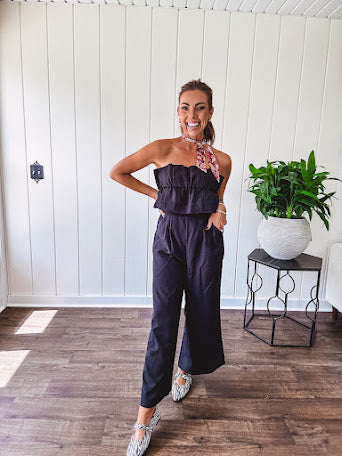 Fully lined black jumpsuit with sleeveless, ruffled bodice, smocked back, wide leg design and pockets at the pleated front.
