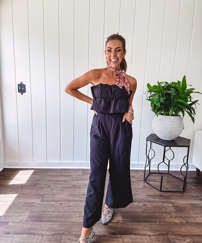 Fully lined black jumpsuit with sleeveless, ruffled bodice, smocked back, wide leg design and pockets at the pleated front.