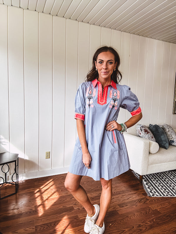 Slate blue dress with puffed, cuffed short sleeves, pockets, a contrasting coral collar and V neck, cuffs and trim and coral and pink embroidery at the yoke.