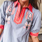 Slate blue dress with puffed, cuffed short sleeves, pockets, a contrasting coral collar and V neck, cuffs and trim and coral and pink embroidery at the yoke.