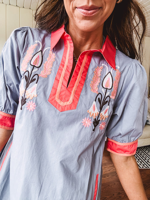 Slate blue dress with puffed, cuffed short sleeves, pockets, a contrasting coral collar and V neck, cuffs and trim and coral and pink embroidery at the yoke.