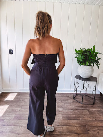 Fully lined black jumpsuit with sleeveless, ruffled bodice, smocked back, wide leg design and pockets at the pleated front.