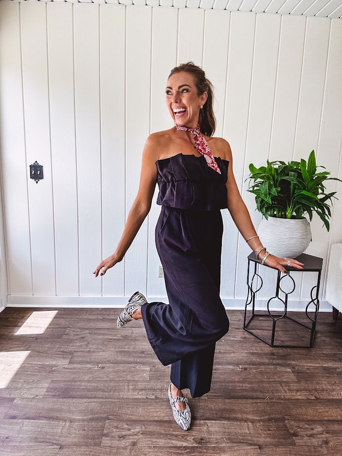 Fully lined black jumpsuit with sleeveless, ruffled bodice, smocked back, wide leg design and pockets at the pleated front.