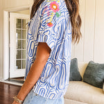 Light blue and white geometric short sleeved button down with collar and cuffed sleeves with bold orange, green and pink embroidery on the shoulders.