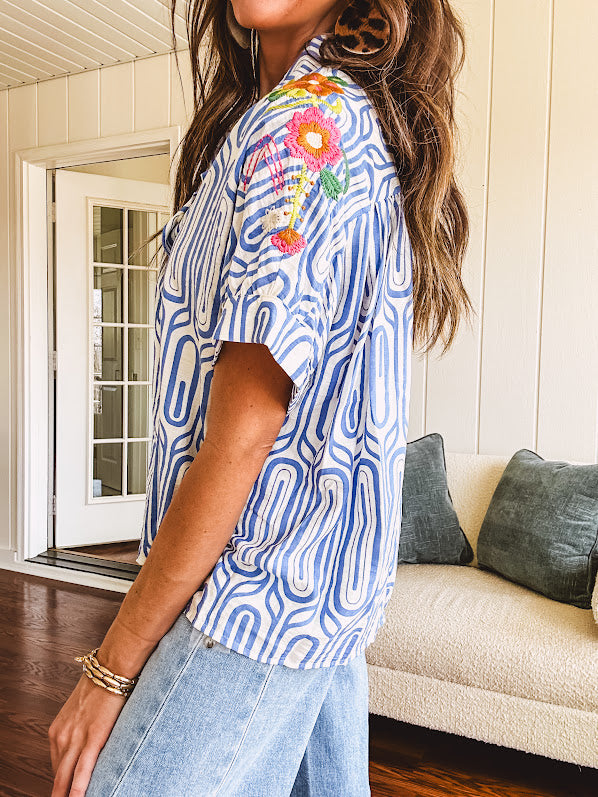 Light blue and white geometric short sleeved button down with collar and cuffed sleeves with bold orange, green and pink embroidery on the shoulders.