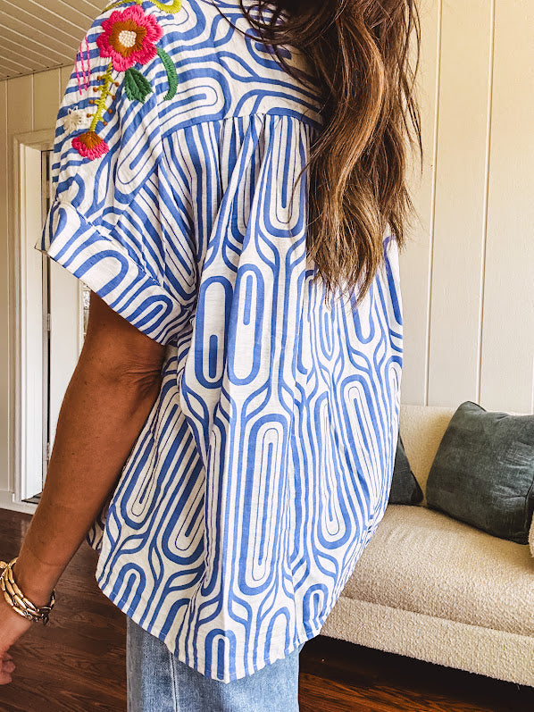 Light blue and white geometric short sleeved button down with collar and cuffed sleeves with bold orange, green and pink embroidery on the shoulders.