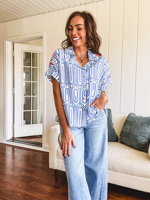 Light blue and white geometric short sleeved button down with collar and cuffed sleeves with bold orange, green and pink embroidery on the shoulders.