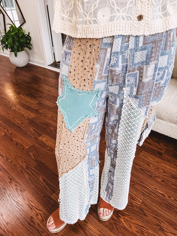 Thin, lightweight wide leg pants with elastic back button and zipper closure, teal, blue and taupe patchwork and mixed fabrics throughout with lace and star patches, pockets and raw hem.