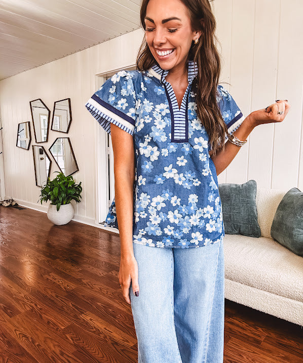 Lightweight cotton denim blue short sleeved top with mock neck and V neck design with shades of blue florals throughout and contrasting navy, white and light blue striping at the neckline and cuffs.