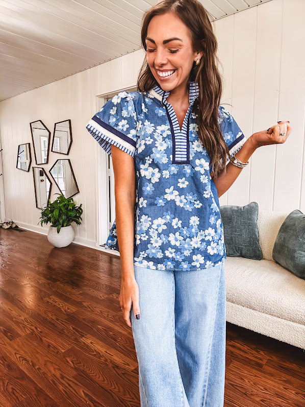 Lightweight cotton denim blue short sleeved top with mock neck and V neck design with shades of blue florals throughout and contrasting navy, white and light blue striping at the neckline and cuffs.