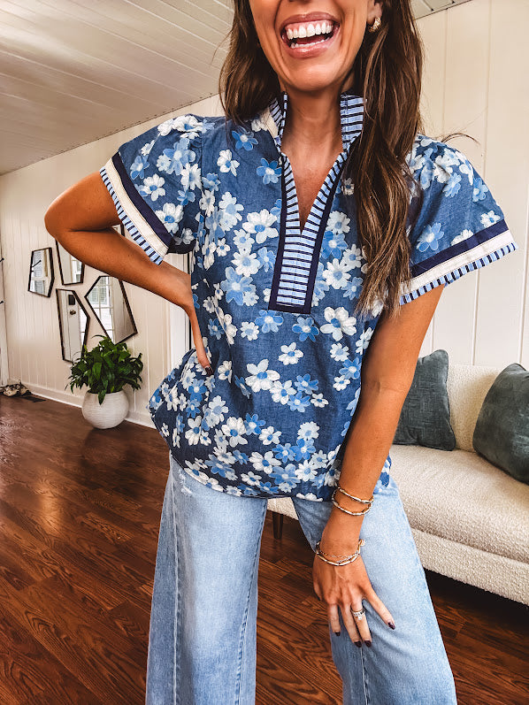 Lightweight cotton denim blue short sleeved top with mock neck and V neck design with shades of blue florals throughout and contrasting navy, white and light blue striping at the neckline and cuffs.
