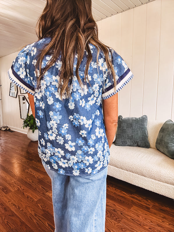 Lightweight cotton denim blue short sleeved top with mock neck and V neck design with shades of blue florals throughout and contrasting navy, white and light blue striping at the neckline and cuffs.