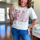 Cream cotton tee featuring "He is Risen" Matthew 28:6 in pink Easter graphics sprinkled with rhinestones.