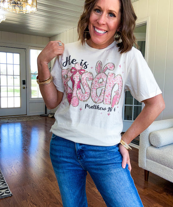 Cream cotton tee featuring "He is Risen" Matthew 28:6 in pink Easter graphics sprinkled with rhinestones.