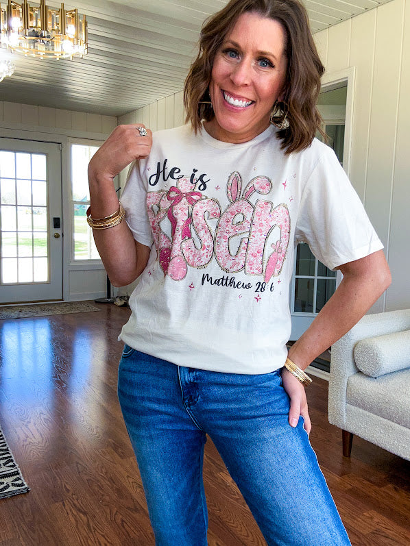 Cream cotton tee featuring "He is Risen" Matthew 28:6 in pink Easter graphics sprinkled with rhinestones.