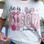 Cream cotton tee featuring "He is Risen" Matthew 28:6 in pink Easter graphics sprinkled with rhinestones.