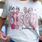 Cream cotton tee featuring "He is Risen" Matthew 28:6 in pink Easter graphics sprinkled with rhinestones.