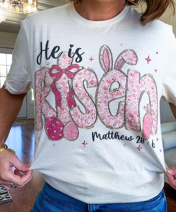 Cream cotton tee featuring "He is Risen" Matthew 28:6 in pink Easter graphics sprinkled with rhinestones.
