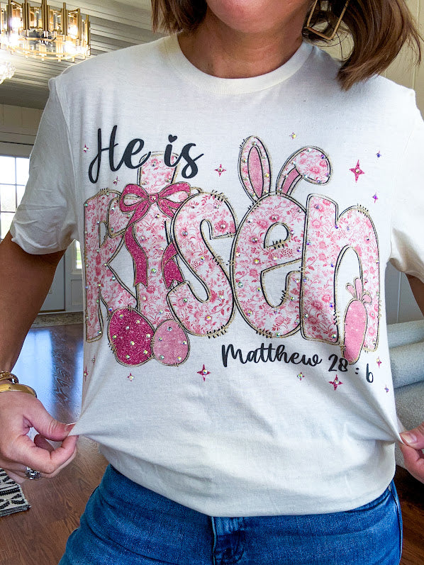 Cream cotton tee featuring "He is Risen" Matthew 28:6 in pink Easter graphics sprinkled with rhinestones.